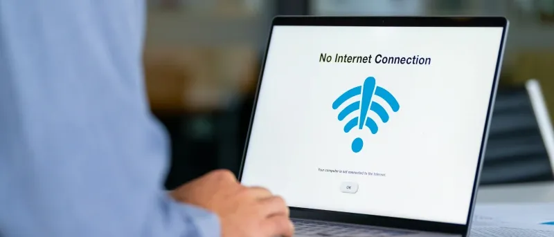 A person wearing a blue shirt is viewed from over the shoulder as they look at a laptop screen displaying a "No Internet Connection" message with a blue Wi-Fi symbol.